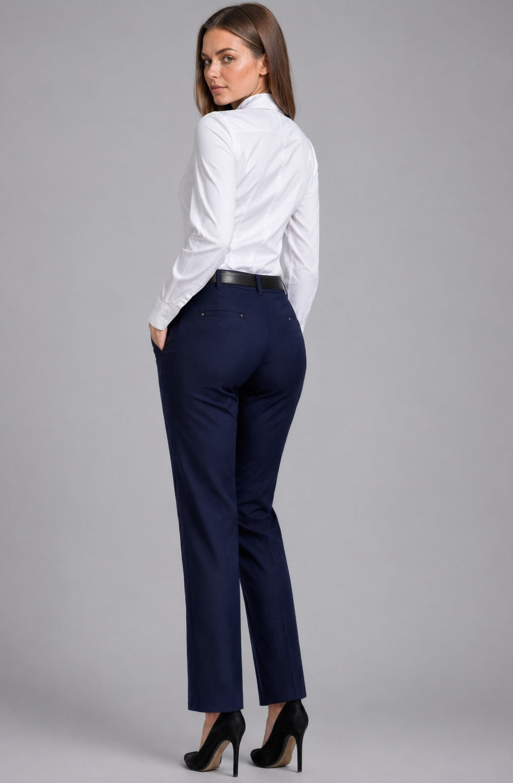 Modelka prezentuje granatowe spodnie damskie MISZTA Atelier z wysokim stanem w ujęciu od tyłu. Elegancki prosty fason z lekko zwężaną nogawką, ozdobnymi patkami z nitami, czarnym paskiem i białą koszulą na jasnym tle studyjnym.